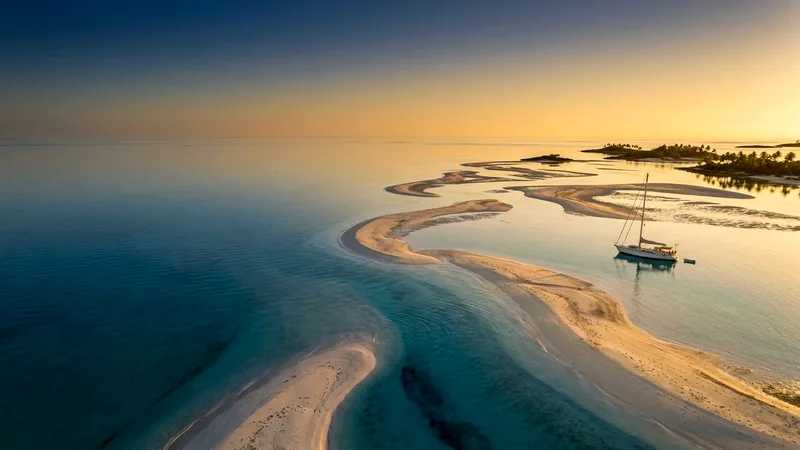 Aerial view of turquoise Exuma Cays sandbars at golden hour with an anchored sailboat.